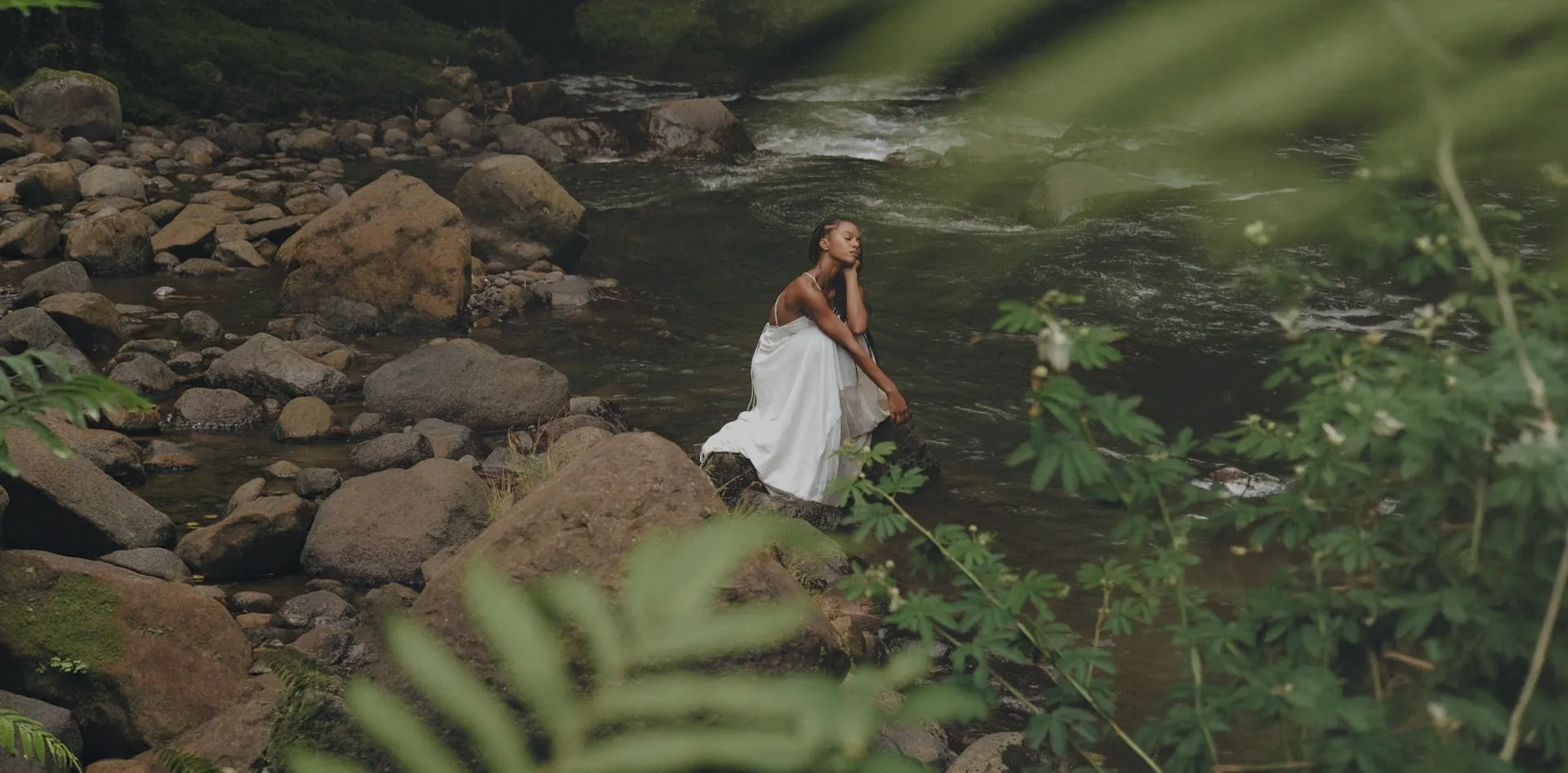 Mujer con vestido blanco sentada sobre una roca en un río del bosque